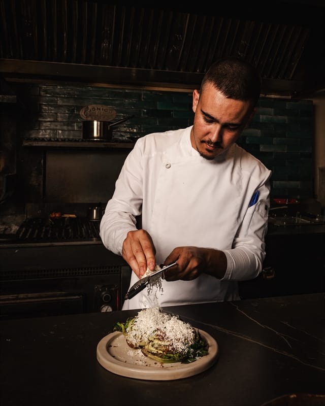 Cocinero con chaquetilla blanca ralla queso sobre una lechuga asada.