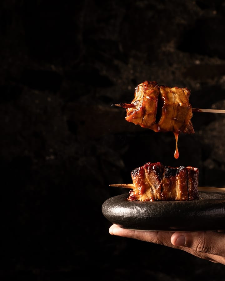 Una brocheta glaseada gotea salsa sobre otra servida en una piedra negra que sostiene una mano.