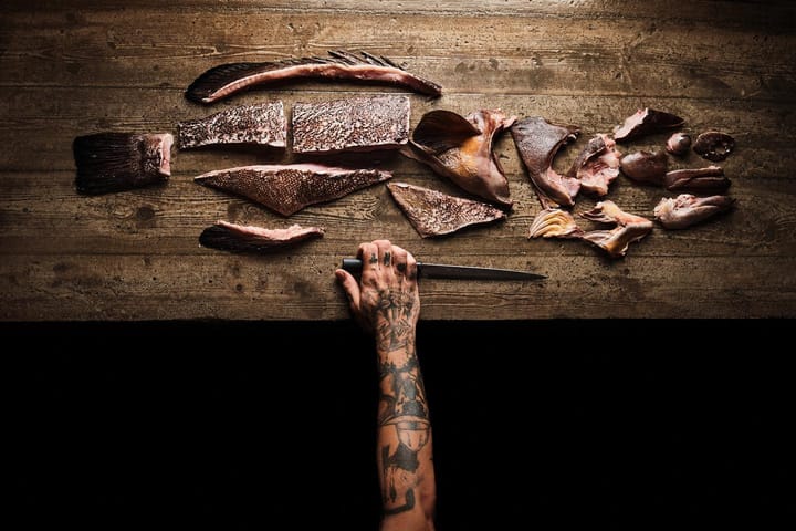 Mano tatuada con un cuchillo junto a un pescado despiezado en lomos, aletas y espinas sobre una mesa de madera.