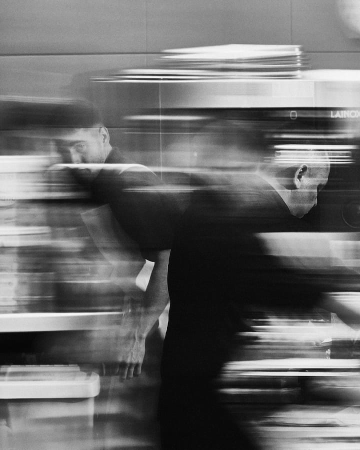 Dos cocineros en el pase, desdibujados por el movimiento; uno mira a cámara entre el acero. En blanco y negro.