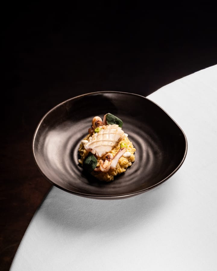 Calamar marcado sobre orzo cremoso con guisantes y hojas, en un plato hondo negro sobre mantel blanco.