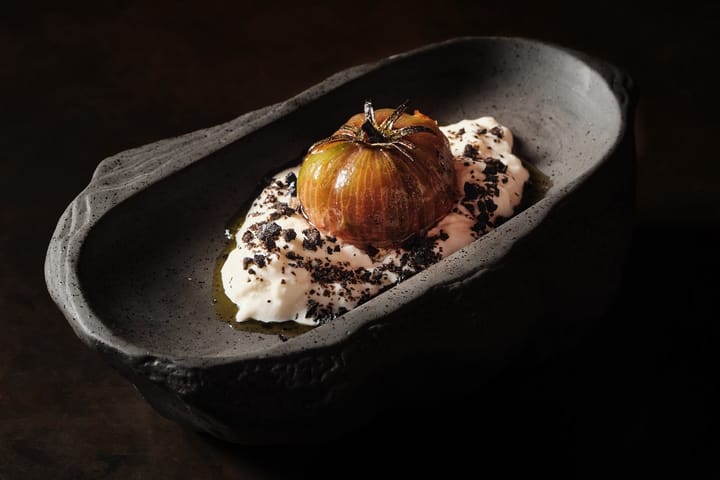 Tomate asado y glaseado sobre una crema blanca con tierra negra, en un cuenco de piedra oscura.