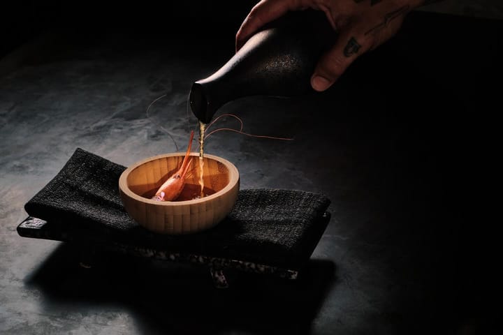 Una mano tatuada vierte caldo desde una botella negra sobre una gamba roja en un cuenco de bambú.