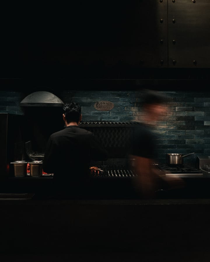 Un cocinero de espaldas trabaja en la parrilla de brasa mientras otro cruza la cocina, desdibujado por la prisa.