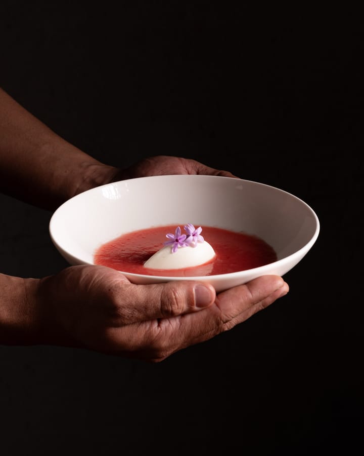 Manos que ofrecen un plato hondo blanco con sopa de fresa, una quenelle blanca y dos flores lilas.