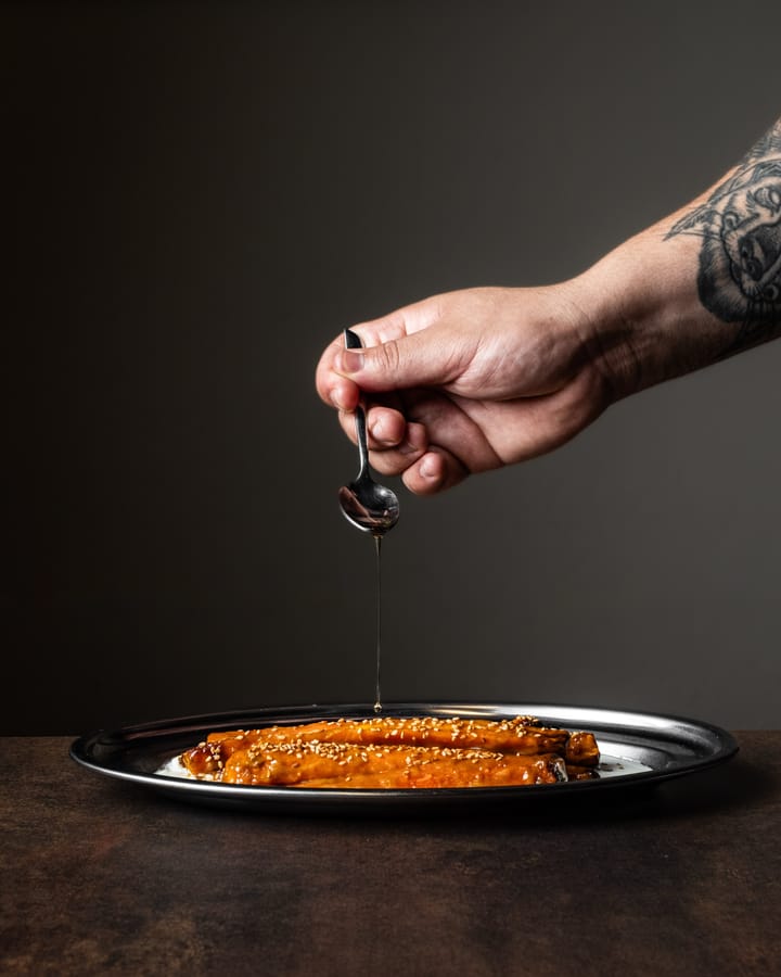 Una mano tatuada deja caer un hilo de aceite con una cuchara sobre zanahorias glaseadas con sésamo.